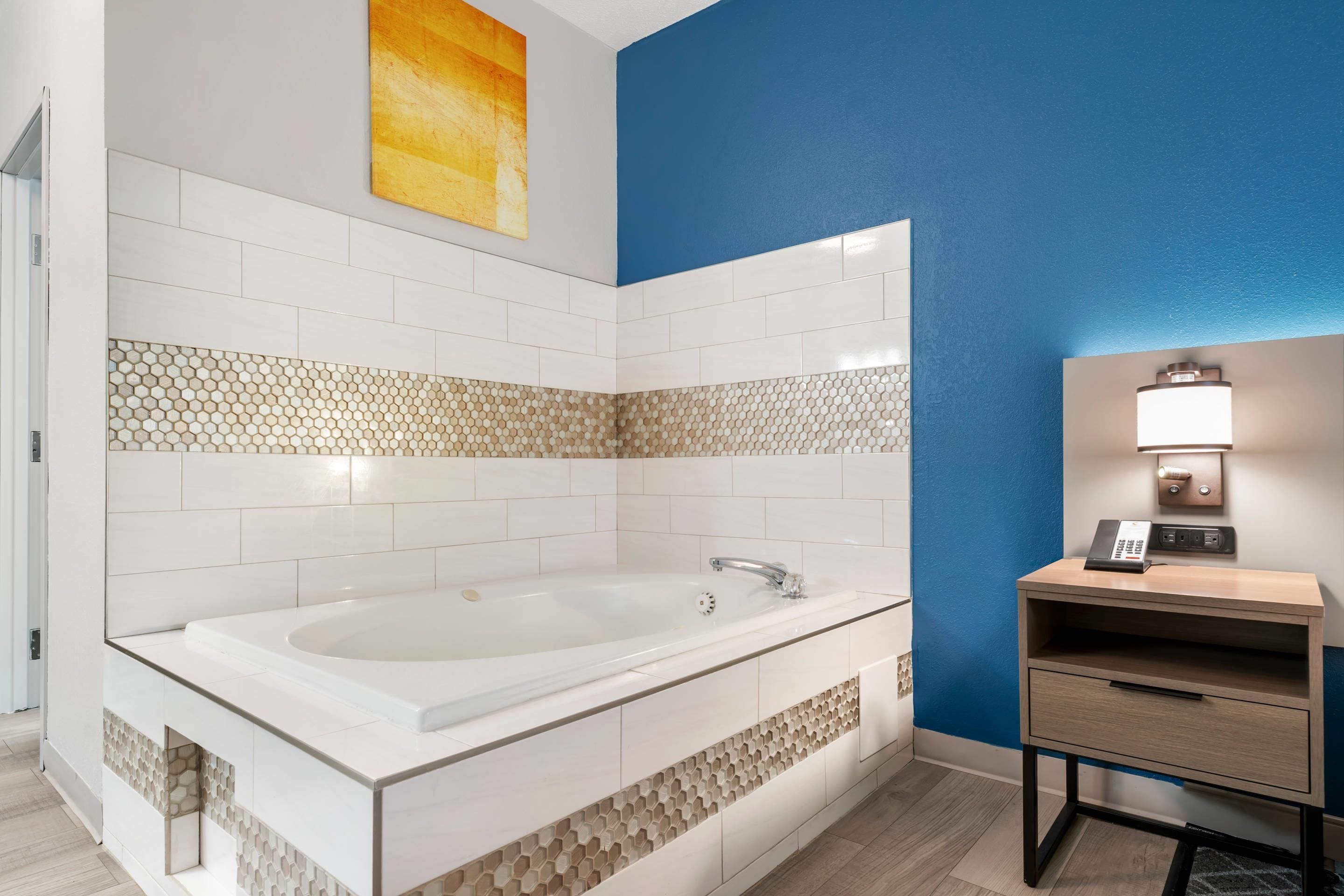 Une salle de bain moderne avec une grande baignoire, des murs carrelés en blanc et en beige, un mur d’accent bleu et une table de chevet avec une lampe.
