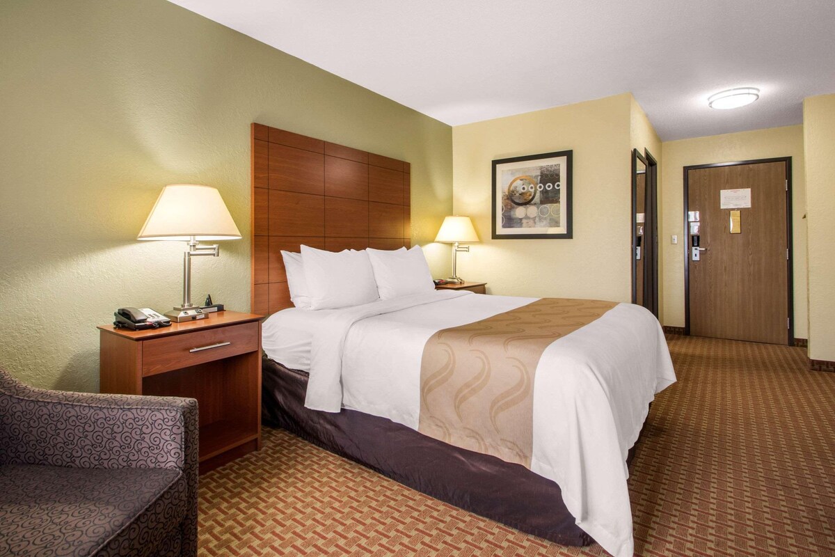 Een hotelkamer met een bed, twee nachtkastjes, een stoel, een lamp, een ingelijst kunstwerk en een deur.