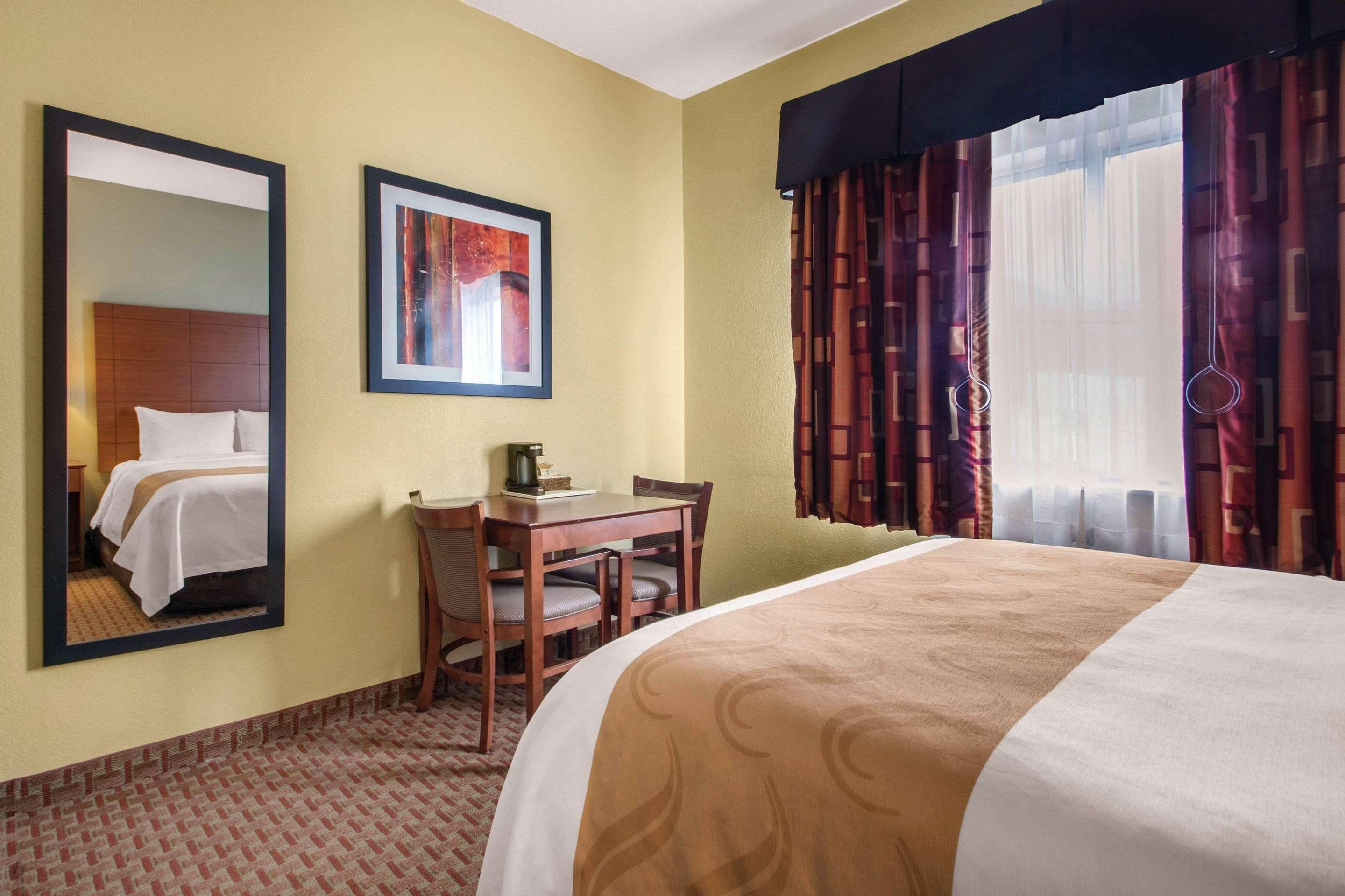 A hotel room with a bed, a desk, a chair, a mirror, a framed picture, and a window with curtains.