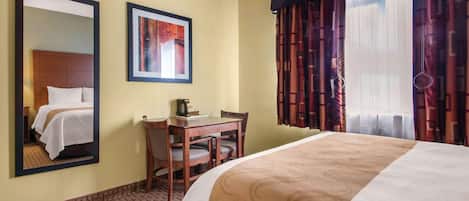 A hotel room with a bed, a desk, a chair, a mirror, a framed picture, and a window with curtains.
