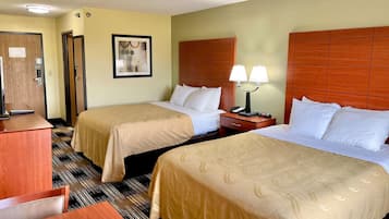 A hotel room with two beds, a desk, a chair, a nightstand with a lamp, and a door.