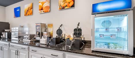 A hotel kitchen with a self-service breakfast bar, waffle maker, coffee machine, and a refrigerator stocked with food items.