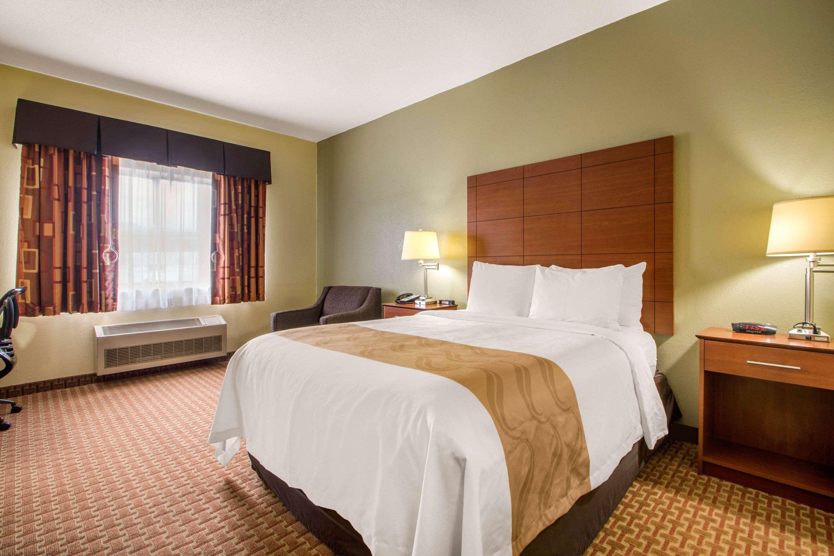 A hotel room with a large bed, two bedside tables, a desk, a chair, a desk lamp, a clock, and a window with curtains.