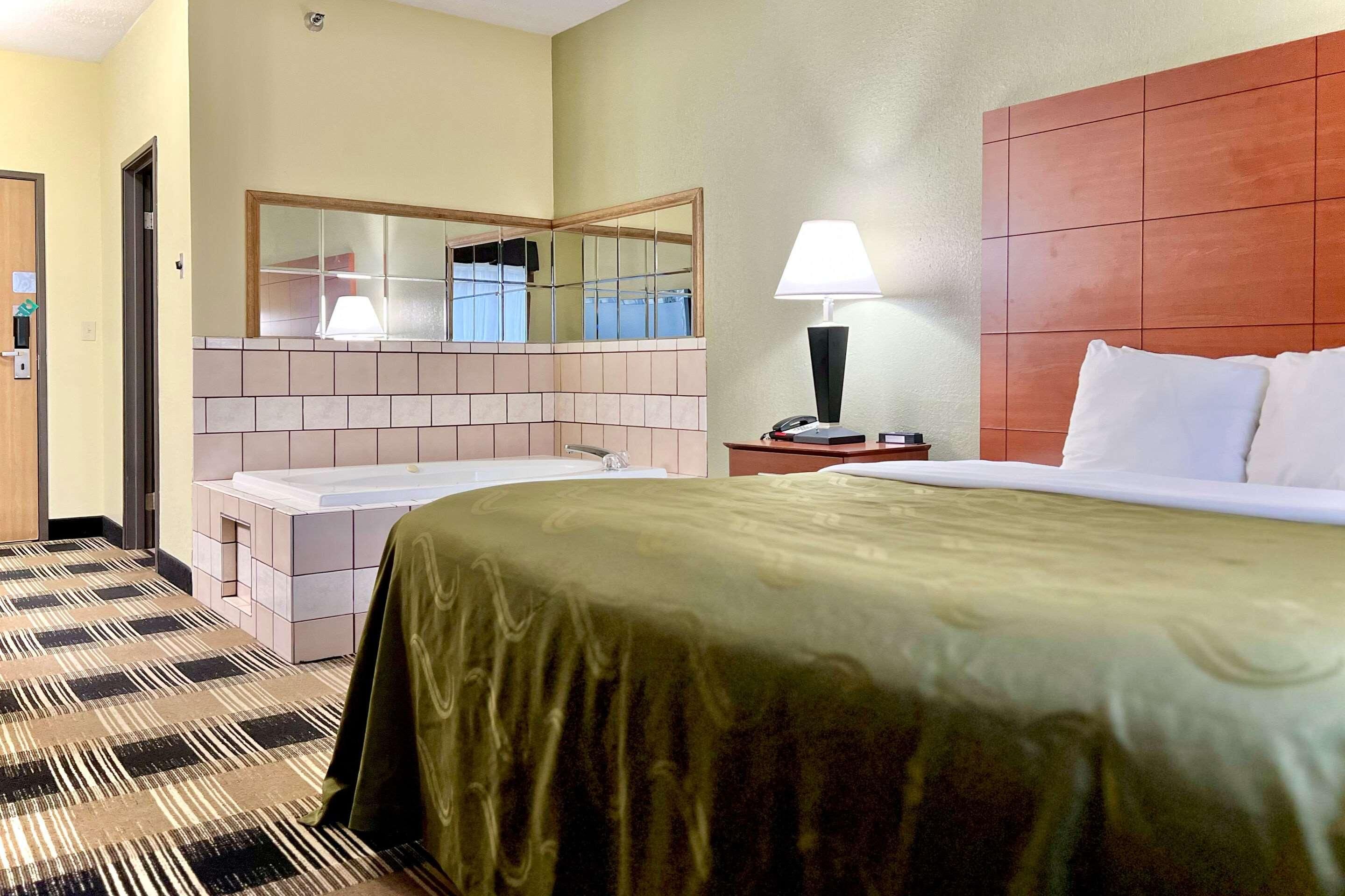 A hotel room with a bed, a bathroom with a sink and mirror, a nightstand with a lamp, and a door.
