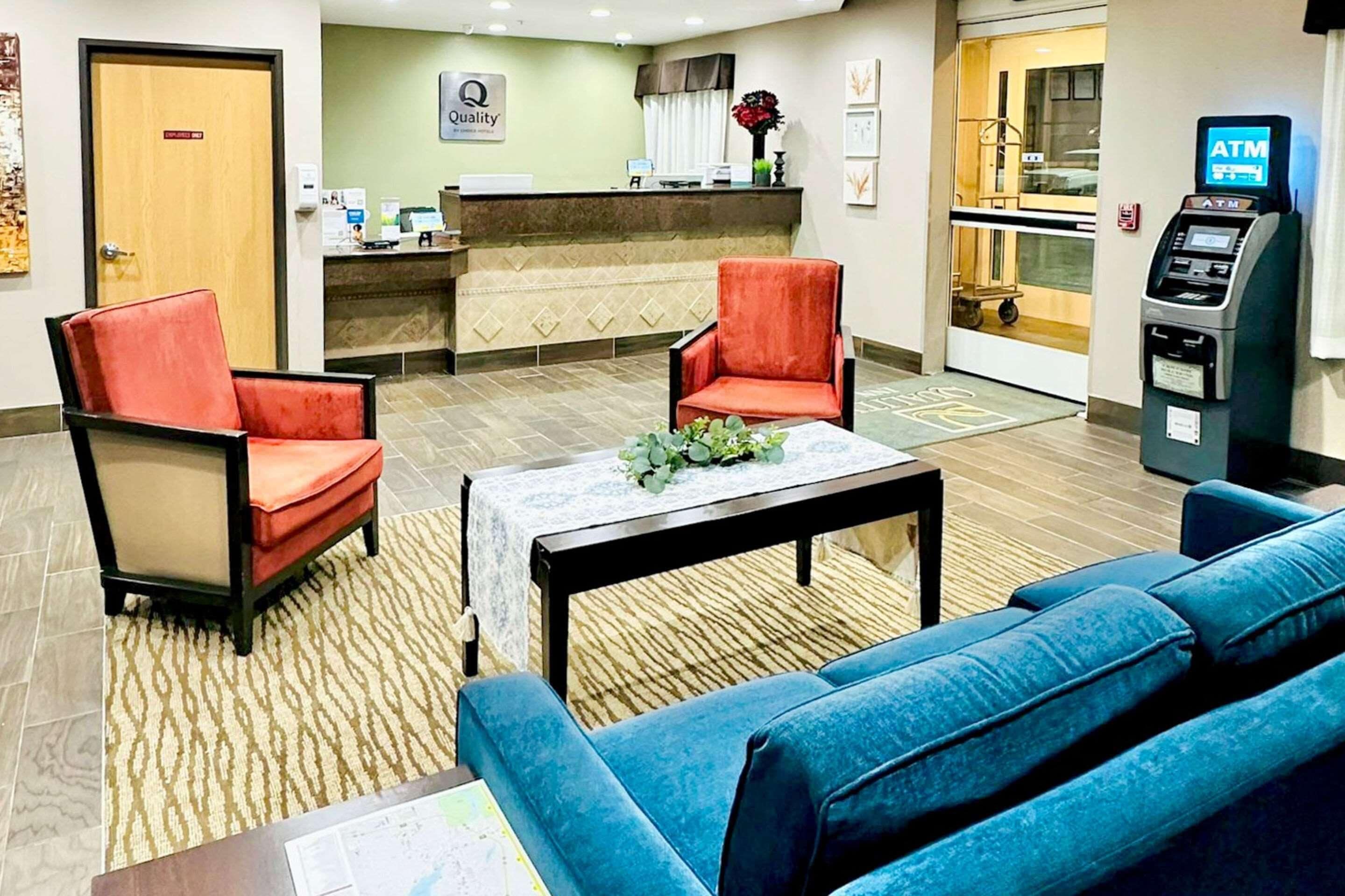 A hotel lobby with a reception desk, two red armchairs, a blue sofa, a small table with a plant, and an ATM machine.