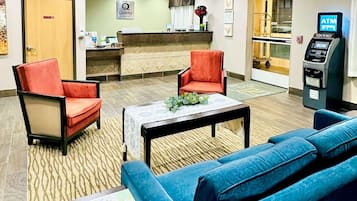 A hotel lobby with a reception desk, two red armchairs, a blue sofa, a small table with a plant, and an ATM machine.