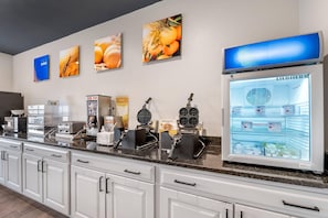 A hotel kitchen with a self-service breakfast bar, waffle maker, coffee machine, and a refrigerator stocked with food items. - Quality Inn & SUI Wisconsin Dells Dwntn-waterparks (Wisconsin Dells)