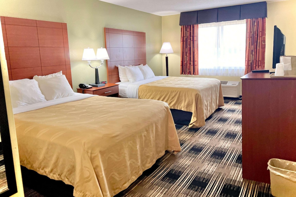 Quarto de hotel com duas camas, uma escrivaninha, uma cadeira, uma televisão e uma janela com cortinas.