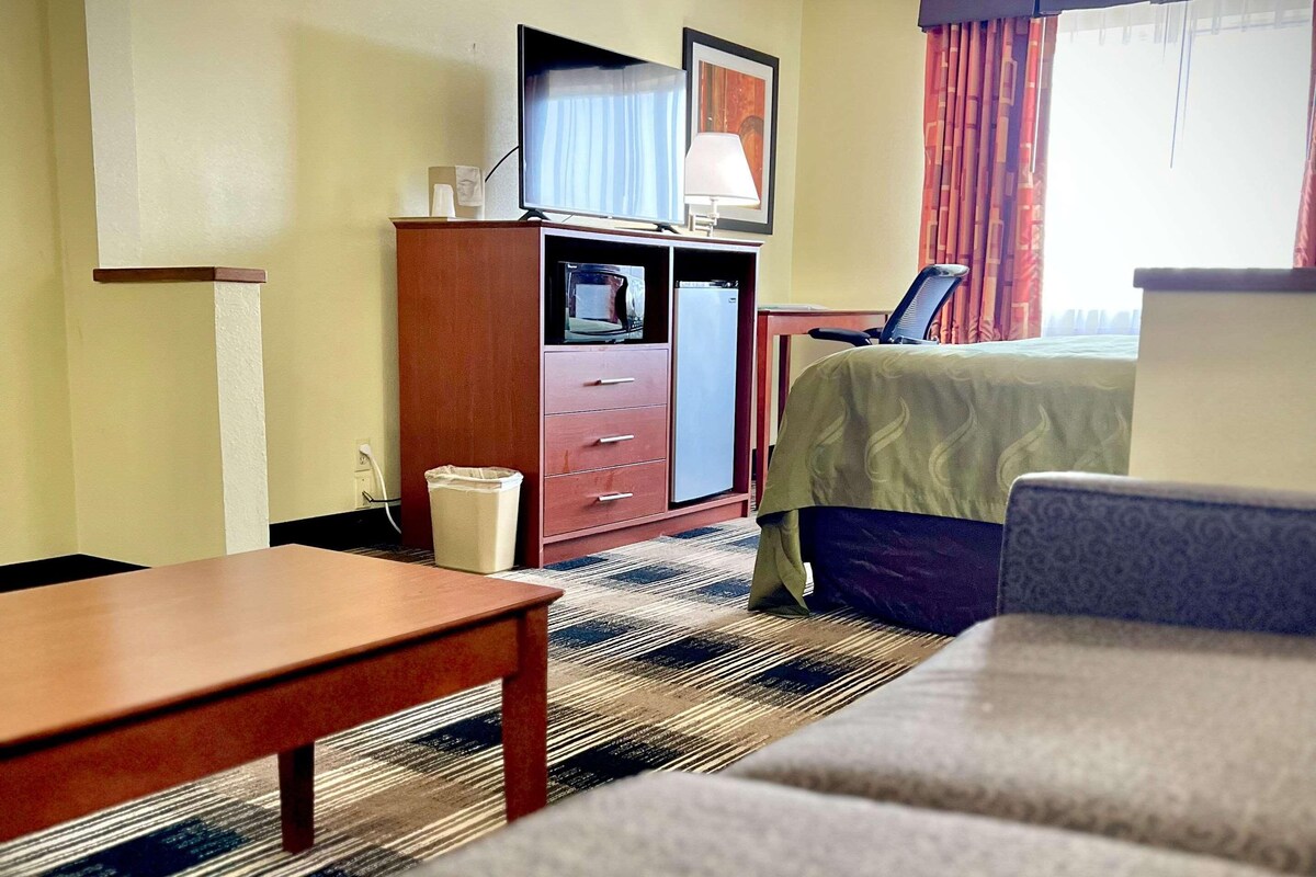Quarto de hotel com cama, escrivaninha, televisão, luminária e cesto de lixo.