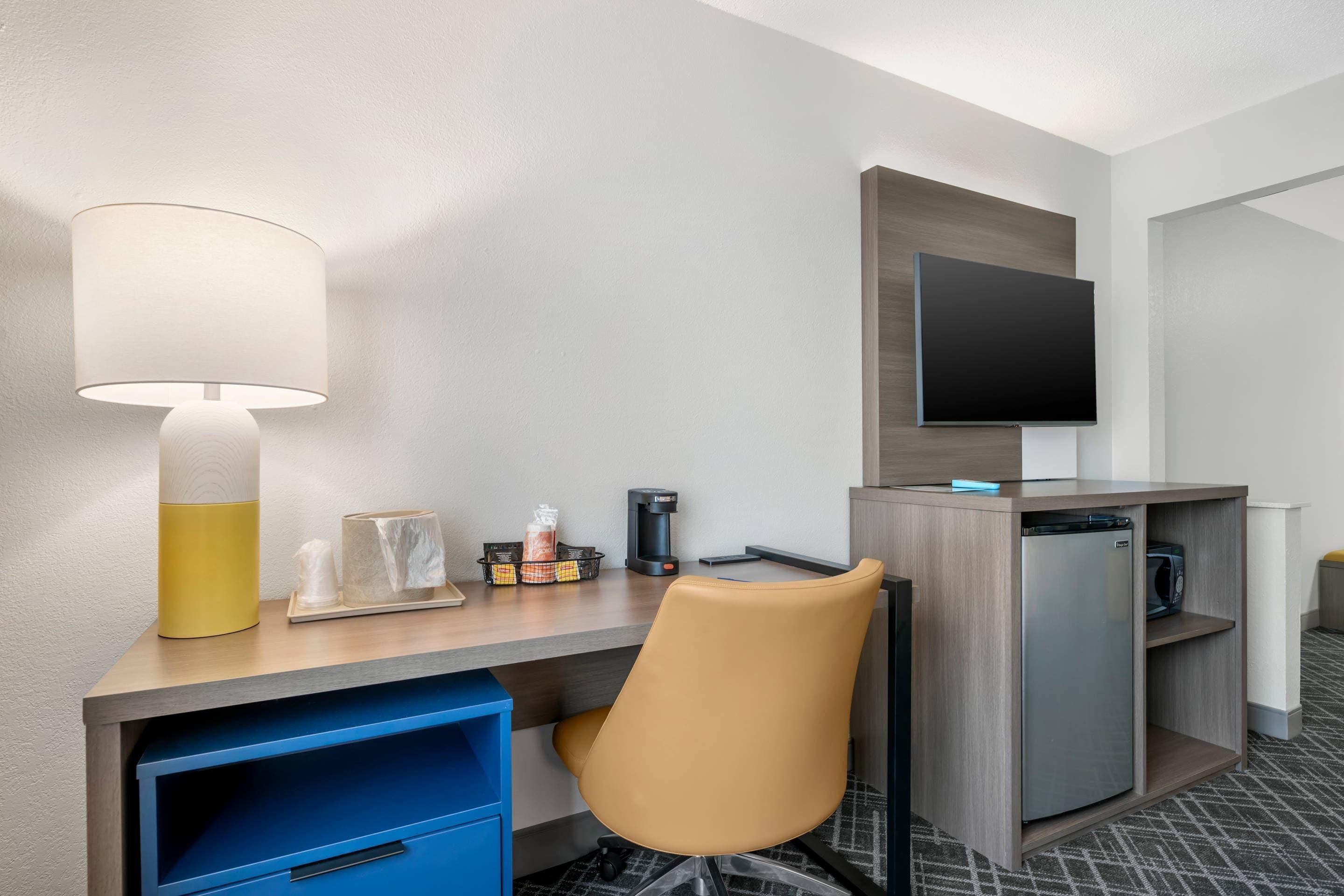 A hotel room with a desk, a chair, a television, a coffee maker, and a refrigerator.