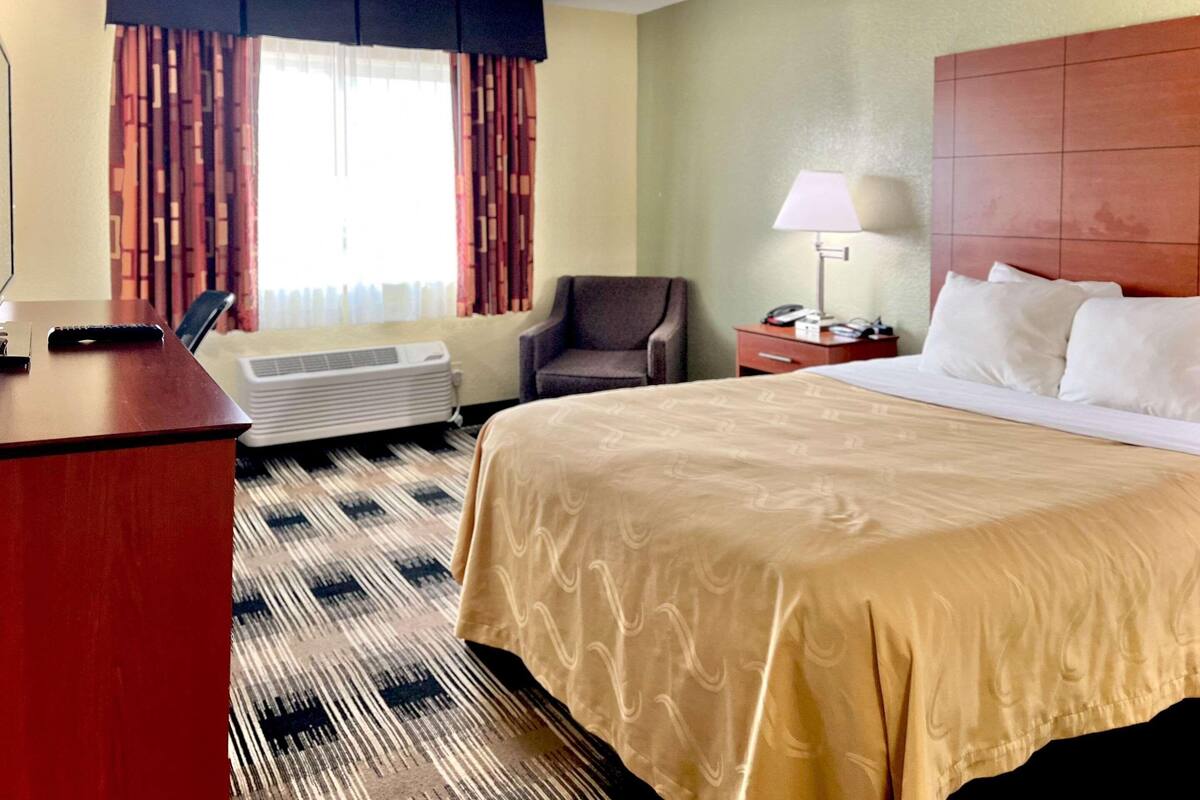 Quarto de hotel com uma cama grande, uma escrivaninha, uma cadeira, um abajur e uma janela com cortinas.