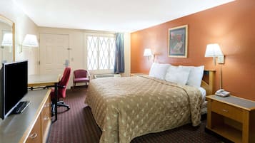A hotel room with a bed, bedside table, lamp, TV, desk, chair, and a window with curtains.