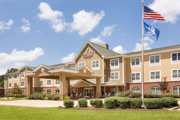 Um hotel com vários andares, fachada bege, entrada central coberta e um mastro com a bandeira americana.