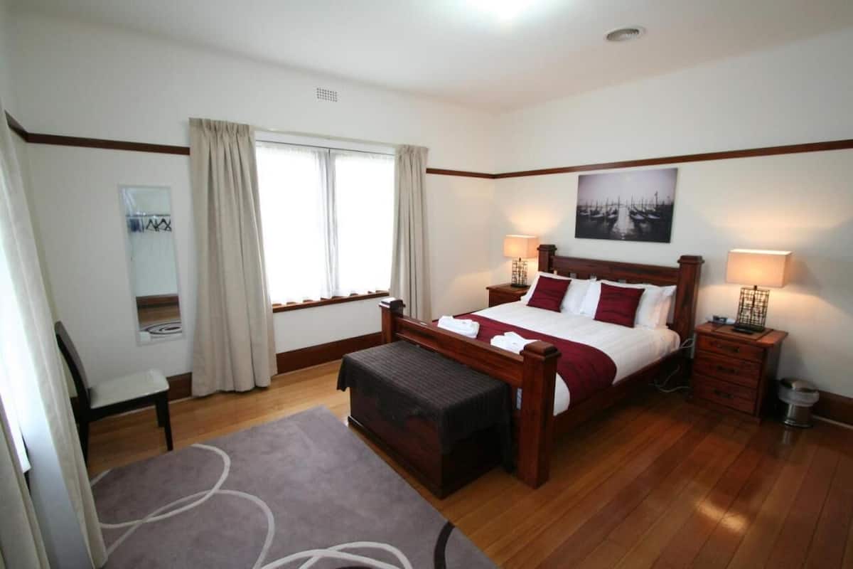 A bedroom with a wooden bed, two bedside tables, a chair, a window with curtains, and a wall-mounted picture.