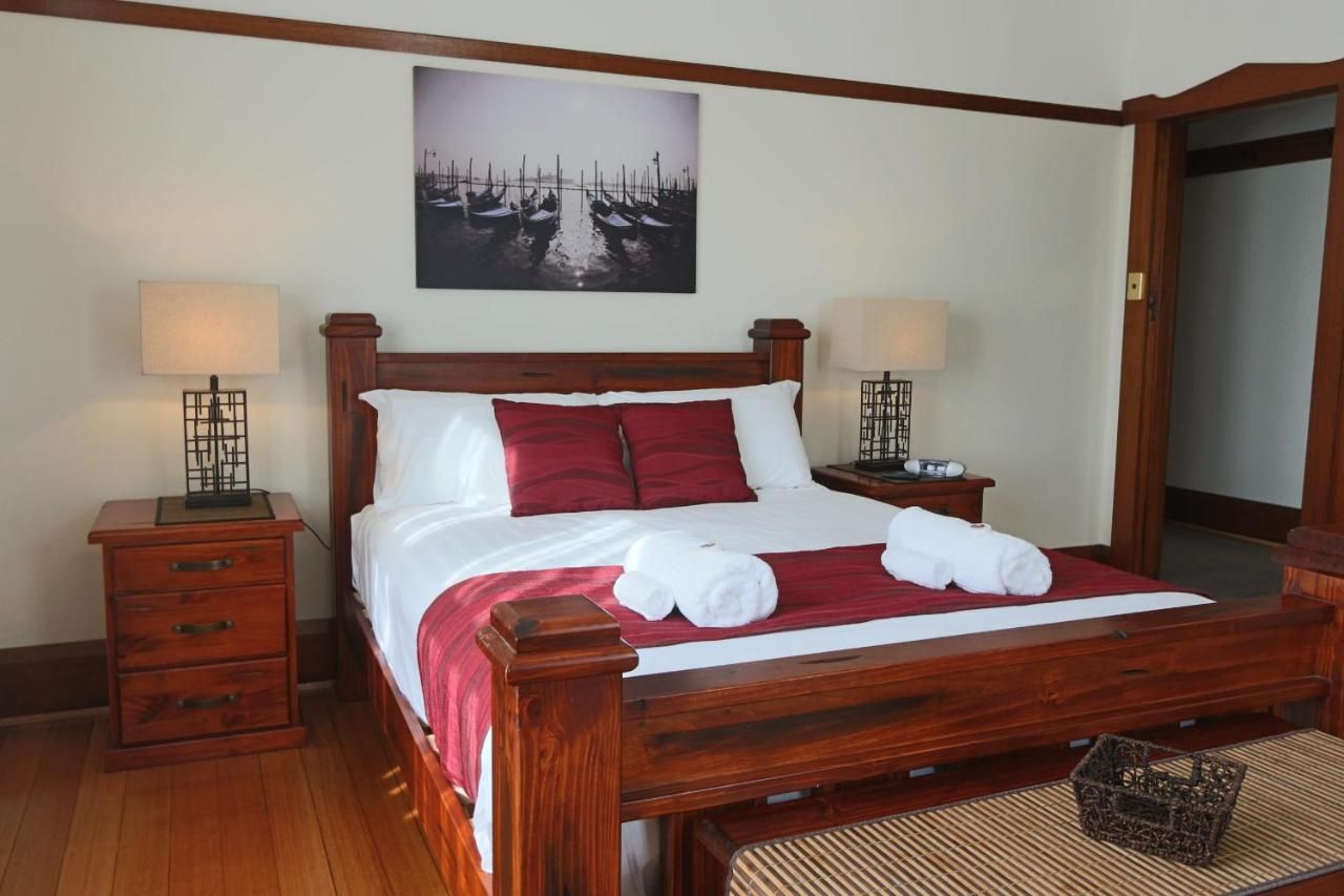 A wooden four-poster bed with white and red bedding, two bedside tables with lamps, a wall-mounted painting, and a woven mat on the floor.