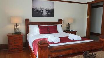 A wooden four-poster bed with white and red bedding, two bedside tables with lamps, a wall-mounted painting, and a woven mat on the floor.