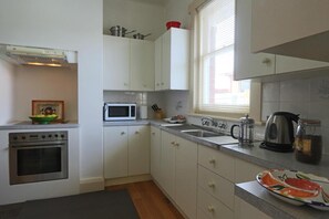 A kitchen with white cabinets, a microwave, a stove, a sink, and a window with blinds. - Moonah Amy Cottage Apartments (Hobart)