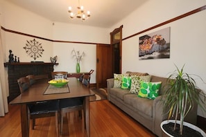 A dining area with a wooden table, chairs, and a vase. There is a sofa with patterned cushions and a potted plant. - Moonah Amy Cottage Apartments (Hobart)