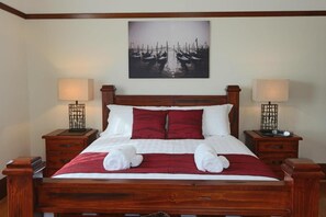 Een netjes opgemaakt bed met een rood-witte dekbedovertrek, twee nachtkastjes met lampen, en een aan de muur hangend schilderij van boten.