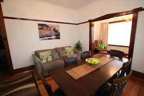 A dining area with a wooden table, chairs, a bowl of fruit, a sofa with patterned cushions, and a painting on the wall. - Moonah Amy Cottage Apartments (Hobart)