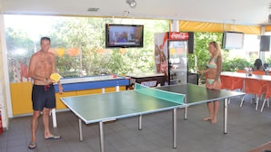 Ett bordtennisbord i ett rum med en tv, en försäljningsautomat för Coca-Cola och orangefärgade stolar.