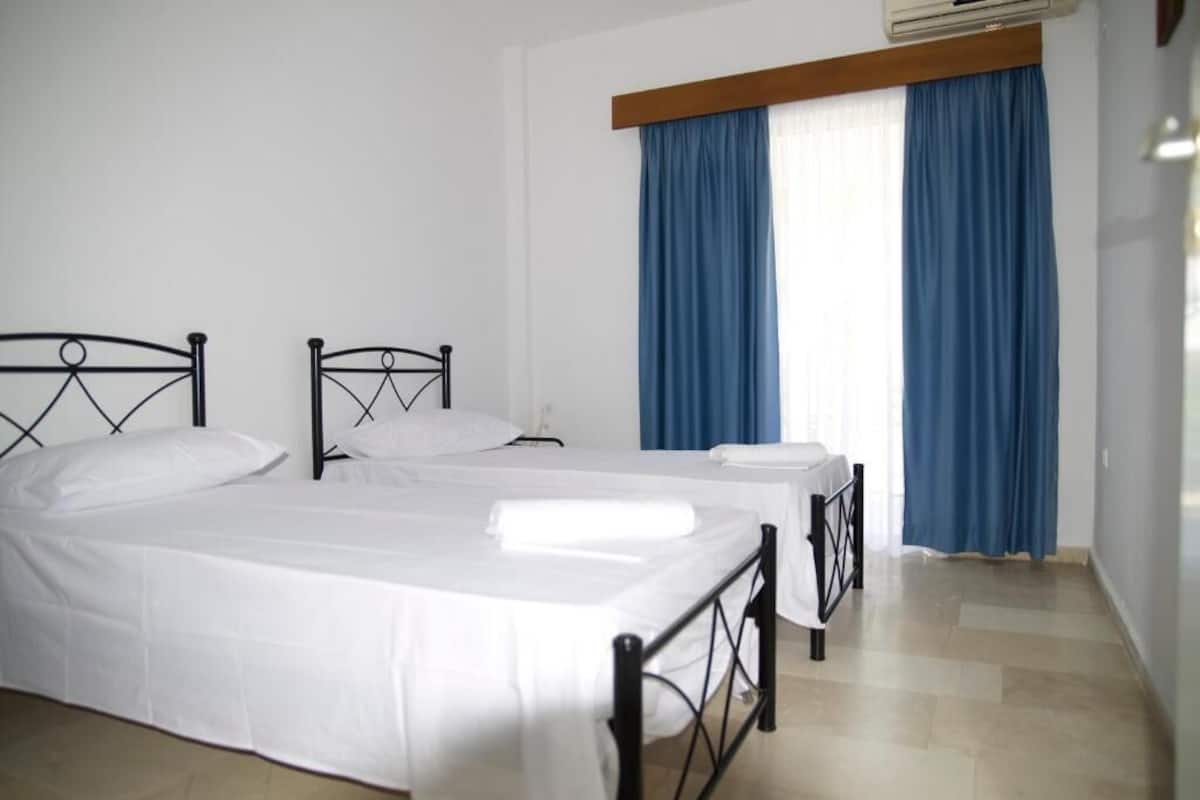 A room with two beds, a window with blue curtains, and an air conditioning unit.