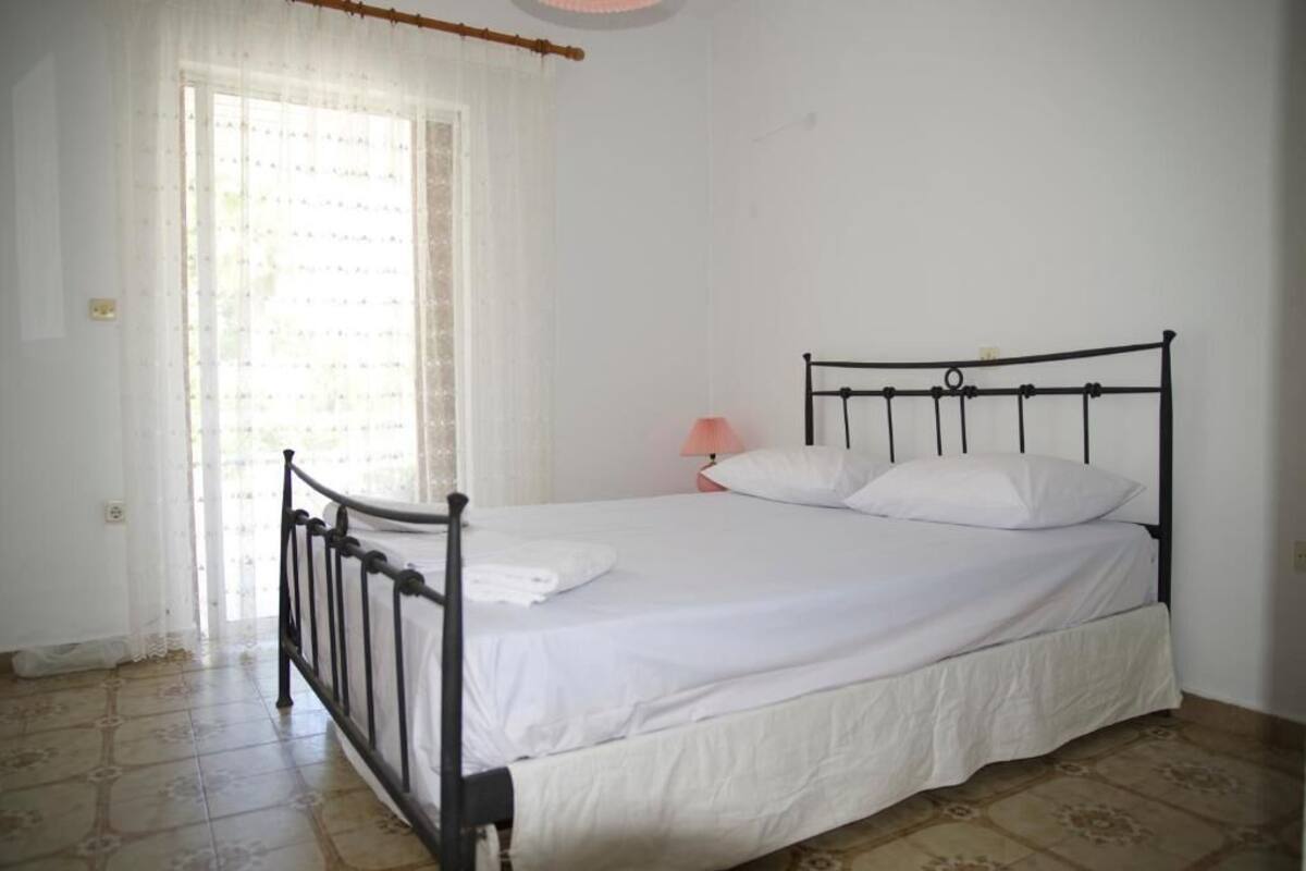 A single bed with a metal frame, white bedding, a pink bedside lamp, and a window with sheer curtains.
