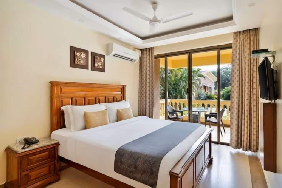 A hotel room with a large bed, a ceiling fan, an air conditioning unit, a balcony with a table and chairs, and a TV mounted on the wall.