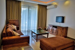 A hotel room with a sofa, ottoman, a small table with a vase, a cabinet, and a flat-screen TV mounted on the wall. - De Mandarin Beach Resort Suites & Villas Goa (Candolim)
