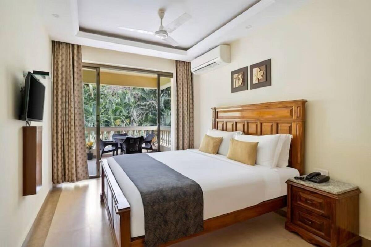 A hotel room with a large bed, a wooden headboard, a bedside table with a phone, a TV mounted on the wall, and a balcony with curtains.