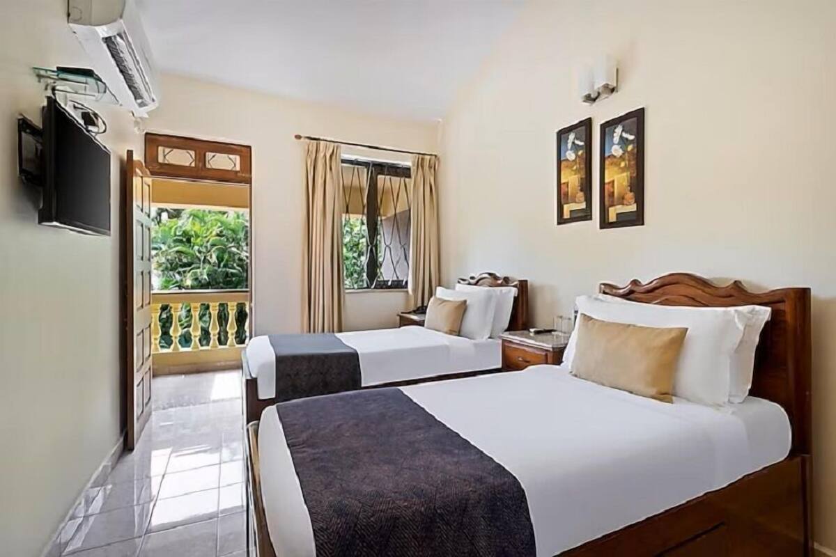 A hotel room with two beds, a TV, a window with curtains, and a balcony with a view of greenery.