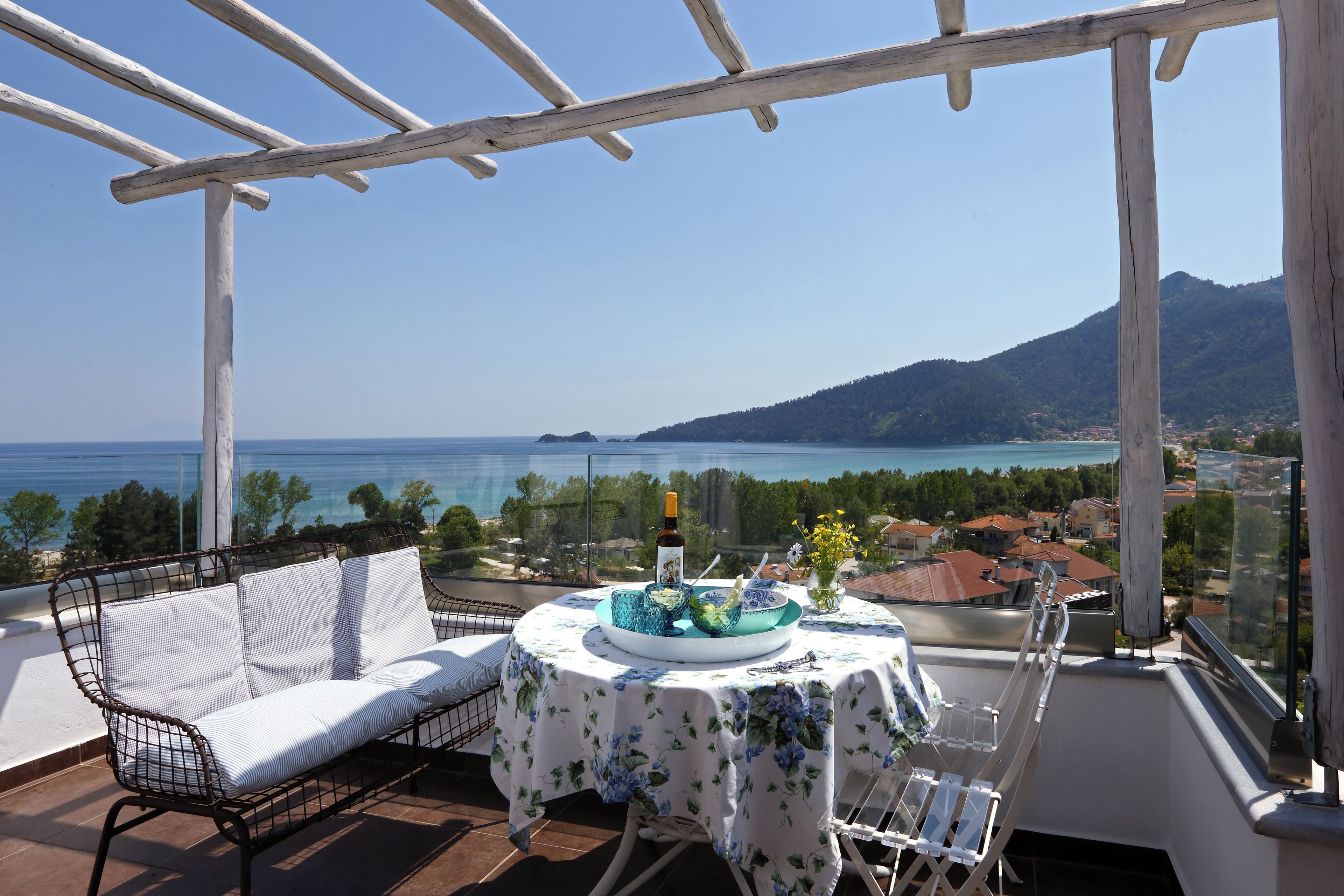 A balcony with a white sofa, a table with a floral tablecloth, and a view of the sea and mountains.