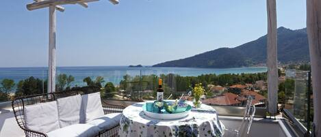 A balcony with a white sofa, a table with a floral tablecloth, and a view of the sea and mountains.