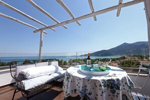 A balcony with a wicker sofa, a table with a floral tablecloth, and a view of the sea and mountains. - Mare Monte Boutique Hotel (Thasos)