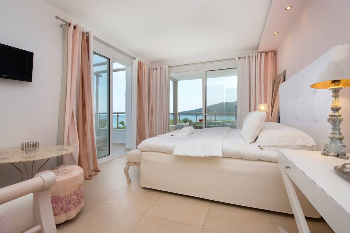 A modern bedroom with a large bed, a desk, a chair, a TV, and a balcony with curtains.