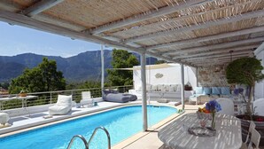 Zona de piscina con vista a montañas, un área de descanso techada y una mesa con un jarrón de flores.