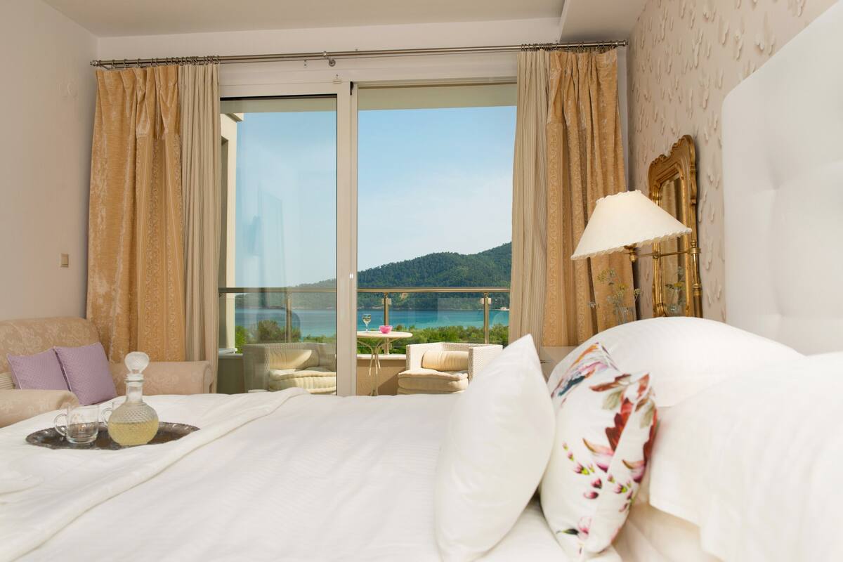 A hotel room with a large bed, a balcony with a view of the sea, and a table with a bottle and glasses.