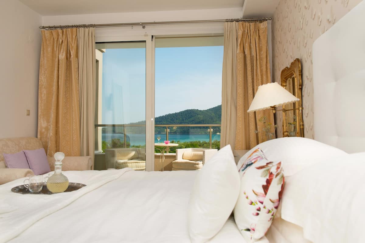 A hotel room with a large bed, a balcony with a view of the sea, and a table with a bottle and glasses.