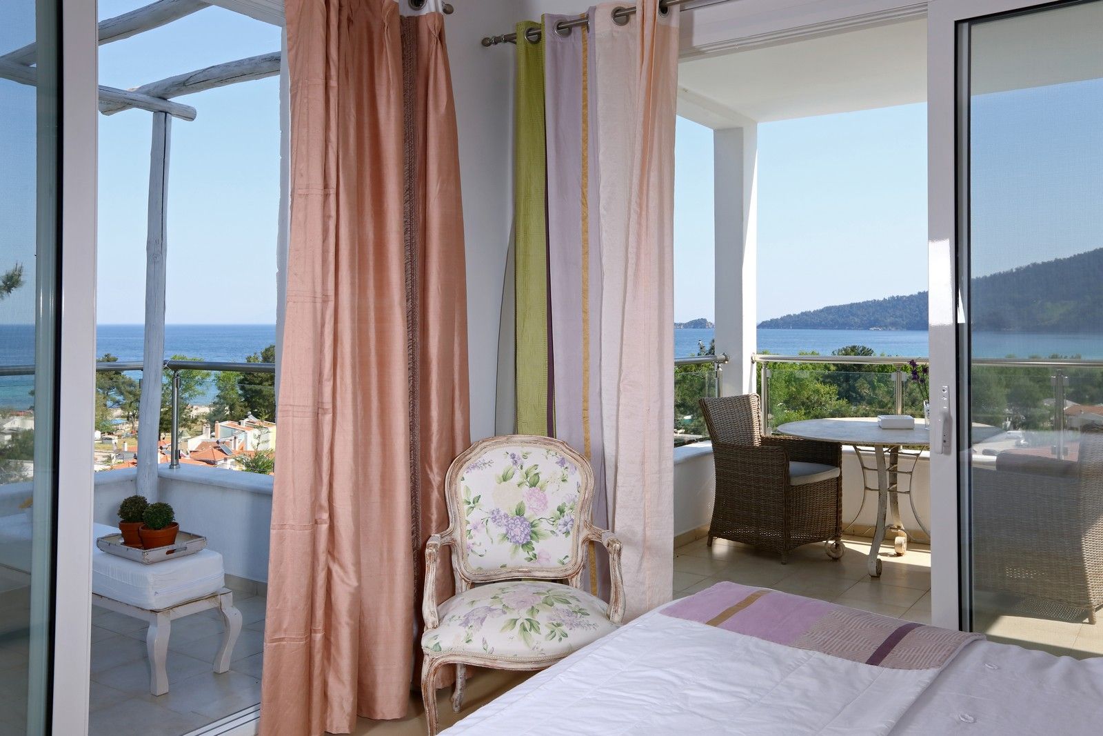 A balcony with a view of the sea, a wicker chair, a small table, and a bed with a view.