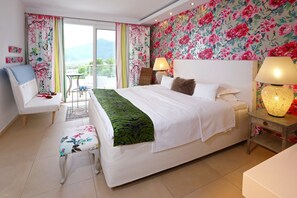 A bedroom with a bed, two bedside lamps, a chair, a small table, and a view of the outdoors. - Mare Monte Boutique Hotel (Thasos)