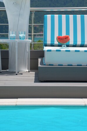 Ein Liegebereich am Pool mit gestreifter Polsterung, einem Glastisch und zwei Gläsern, sowie einer Wassermelonenscheibe auf der Polsterung.