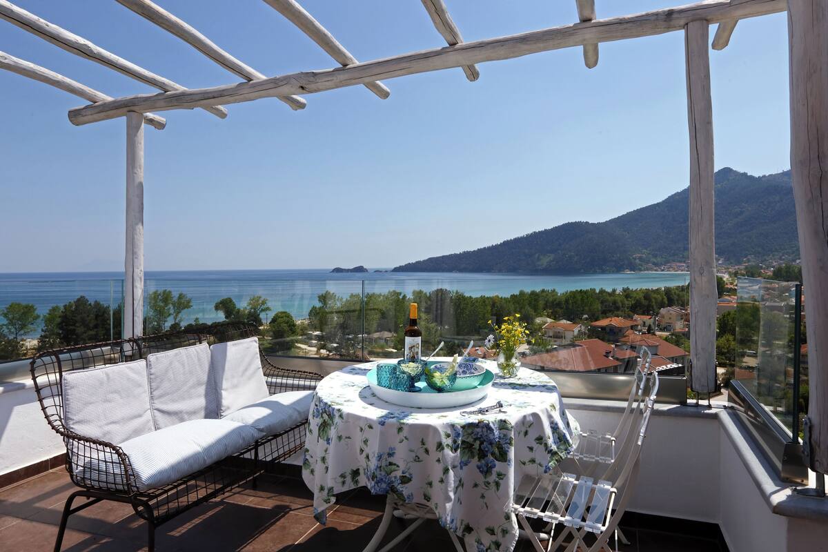 A balcony with a white sofa, a table with a floral tablecloth, and a view of the sea and mountains.