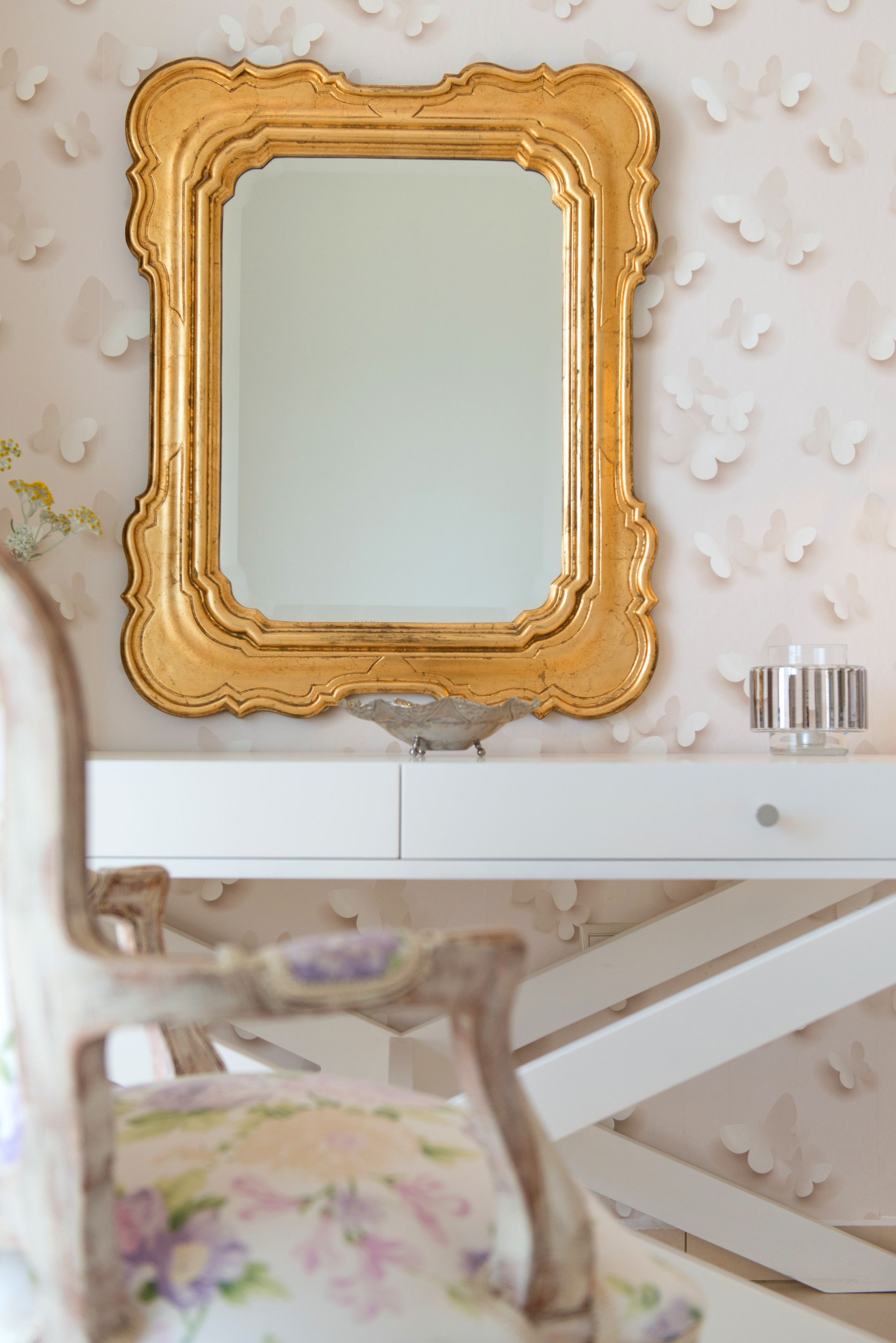 Une coiffeuse blanche avec un miroir encadré d’or, une chaise à motifs floraux et un mur décoratif orné de motifs de papillons.