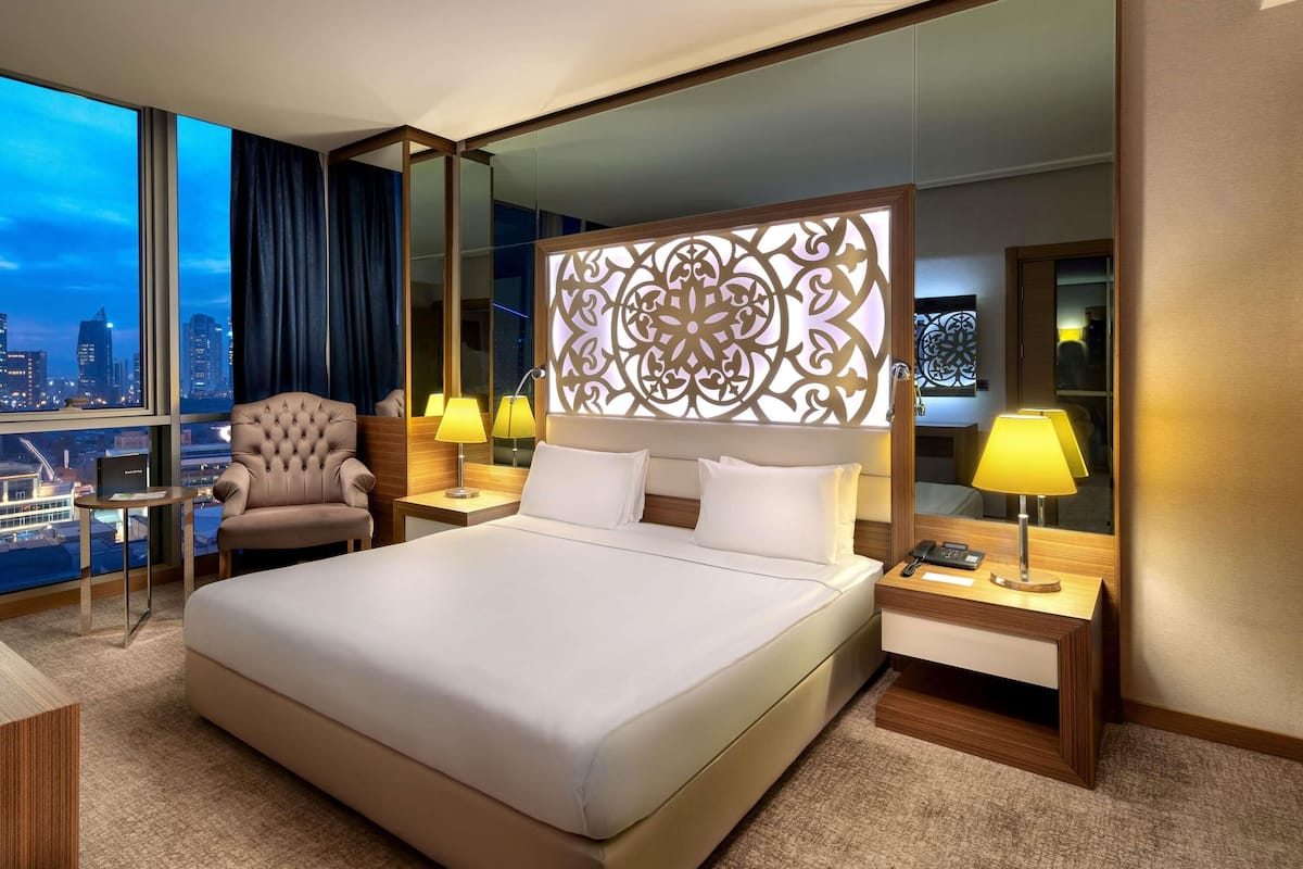 Habitación de hotel con una cama grande, una silla, un escritorio y vistas a la ciudad.