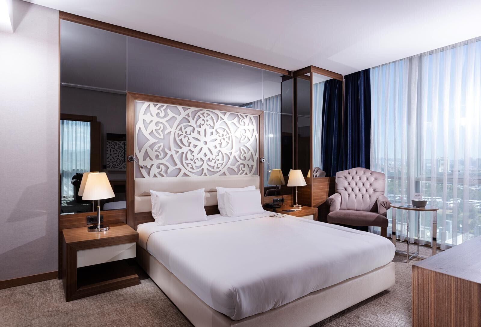A hotel room with a large bed, two bedside tables, a chair, a small table, and a view of the city through large windows.