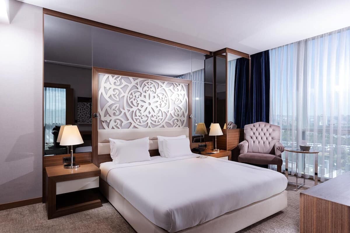 Habitación de hotel con una cama grande, dos mesitas de noche, una silla, una mesita pequeña y vistas a la ciudad a través de amplias ventanas.