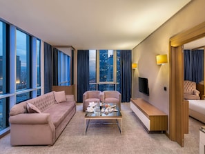 Una habitación de hotel moderna con un amplio ventanal, un sofá, dos sillones, una mesa de centro, un televisor y una mesita de noche.