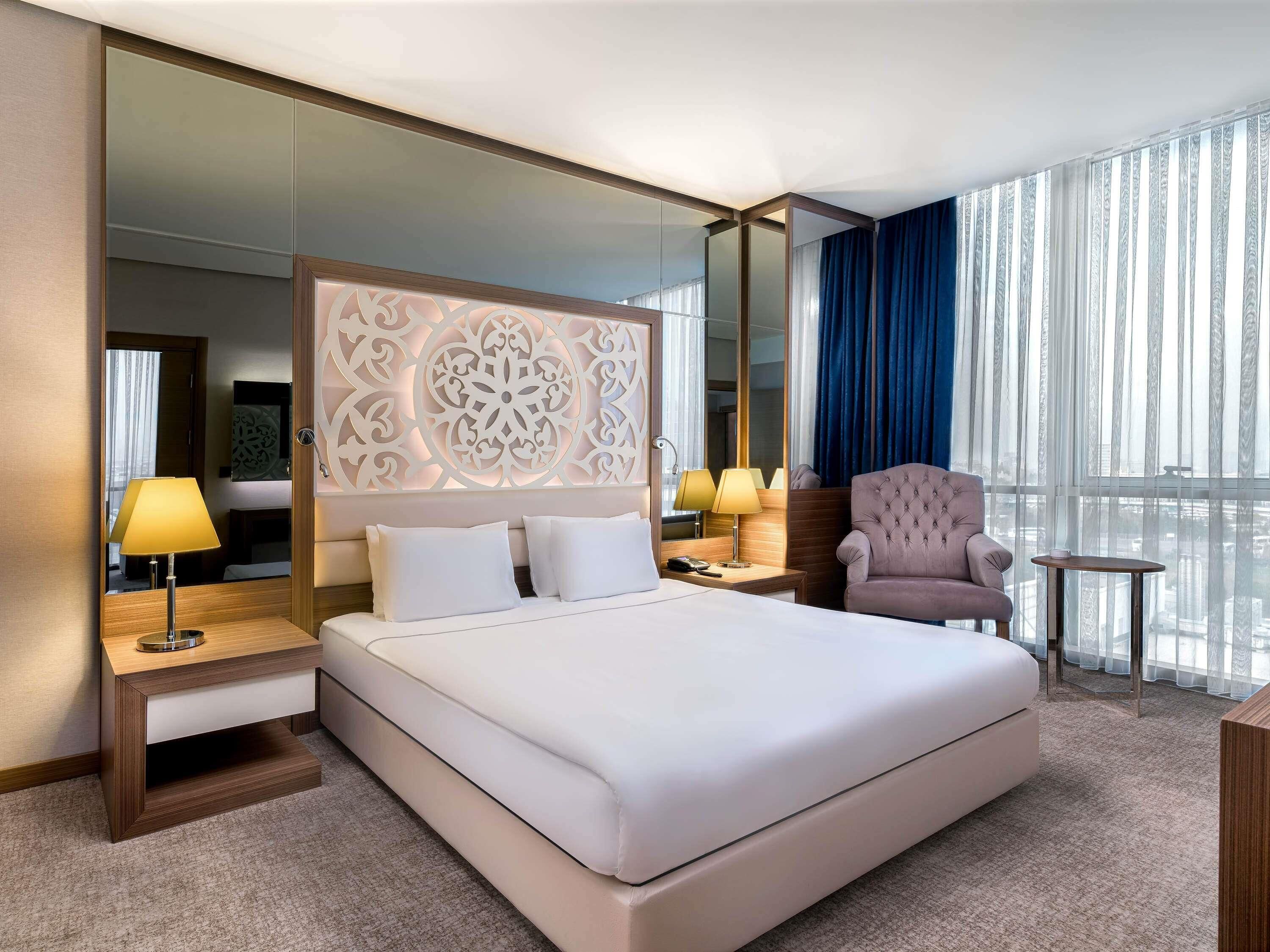 A hotel room with a large bed, two bedside tables, a chair, a small table, and a large window with curtains.