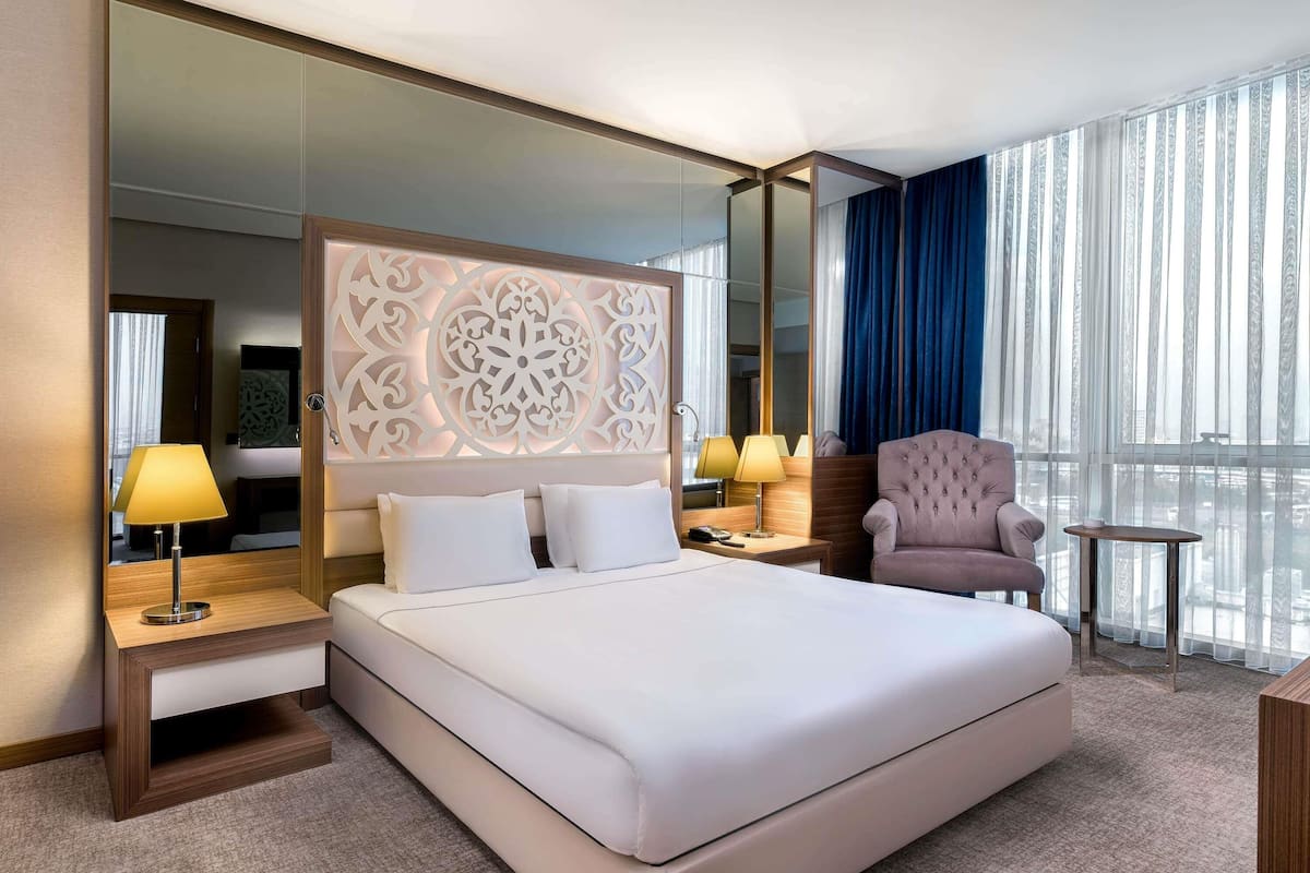 Habitación de hotel con una cama grande, dos mesitas de noche, una silla, una mesita pequeña y un amplio ventanal con cortinas.