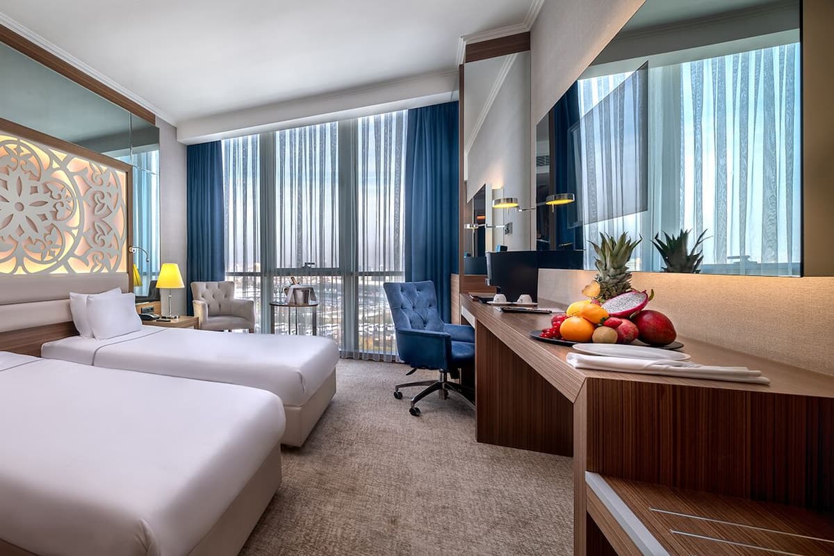 Habitación de hotel con dos camas, un escritorio, una silla, un ventanal grande con cortinas y un panel decorativo en la pared.